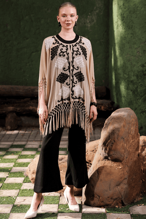 Front view of model wearing beige cape with black and white embellishment and fringe detail