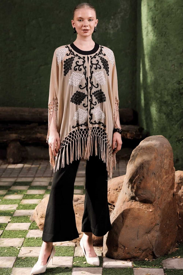 Front view of model wearing beige cape with black and white embellishment and fringe detail